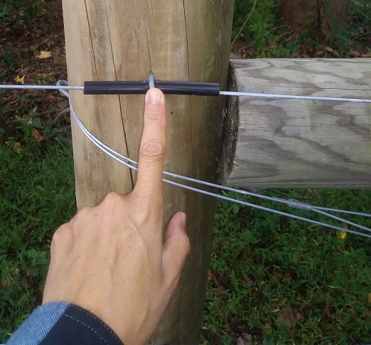 A high tensile fence post with a plastic insulator around an electrified wire. A metal staple holds the insulator to the fence post. I am touching the staple with my finger.
